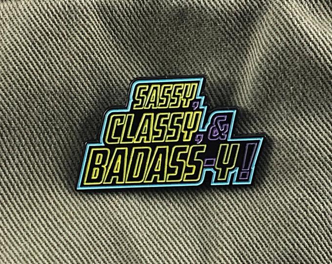A multi-color, empowering enamel pin with bold stacked "Sassy, Classy, & Badass-y!" stylized lettering, channeling retro 80s synthwave, dark-mode vibes in neon blue, purple, yellow hues.